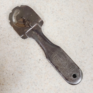 May include: A dark metal vintage can opener featuring a flat handle with an embossed shield logo containing the text ACORN. The tool includes a circular cutting mechanism at the top, a small hole at the base of the handle, and text reading MADE IN U.S.A.