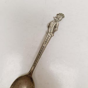 May include: Vintage silver-toned spoon featuring a detailed figure of Woody Woodpecker at the handle's top. The spoon's handle is inscribed with "WOODY WOODPECKER". The spoon has a classic, slightly worn appearance.