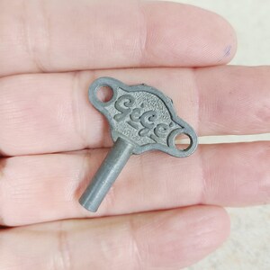 May include: A small, grey metal winding key featuring a textured, wing-shaped top with two circular holes and the embossed text "Gege" in a cursive script. The key has a cylindrical shaft extending from the center of the textured head.