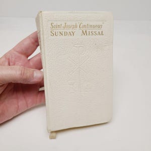 May include: A white, textured hardcover book titled Saint Joseph Continuous Sunday Missal, featuring gold embossed lettering and a Chi-Rho symbol with a chalice design debossed on the front cover, including a small fabric ribbon bookmark at the base.