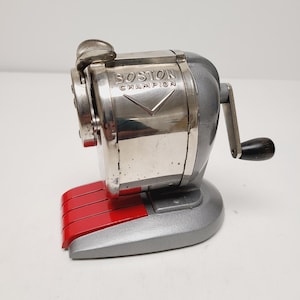May include: A vintage Boston Champion pencil sharpener. The sharpener is silver and gray with a red pencil guide. The words "BOSTON CHAMPION" are printed on the front. It has a crank handle with a black knob.