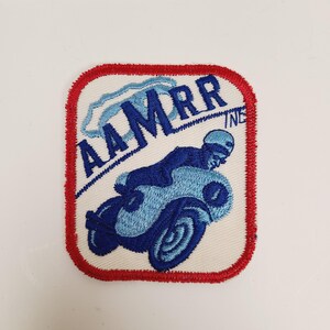 May include: A white embroidered patch with a red border. The patch features a blue motorcycle with a rider and the text "AAMRR INC."