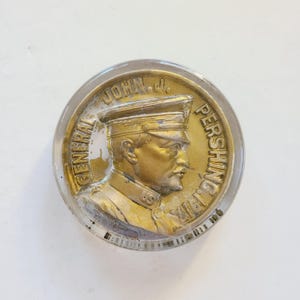 May include: A glass paperweight with a gold-toned metal medallion featuring a profile portrait of General John J. Pershing in military uniform. The medallion is inscribed "GENERAL JOHN J. PERSHING WWI".
