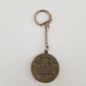 May include: A vintage keychain with a circular bronze-colored pendant. The pendant features an image of a building and the text "1988 Summit of Industrialized Nations". The keychain has a silver-colored ring and chain.