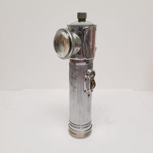 May include: A vintage, chrome-plated flashlight with a ribbed cylindrical body and a large, round lens. The flashlight features a switch and a top cap. The design is reminiscent of early 20th-century lighting technology.