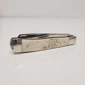 May include: A closed pocket knife with a cream-colored handle and silver-colored metal accents. The handle features a map design and the word "MOORE" is printed on the end. The knife has multiple blades.