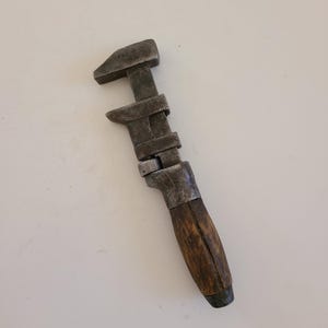 May include: An antique adjustable wrench with a dark, weathered metal finish. The handle is made of worn, dark wood. The wrench is designed for tightening or loosening nuts and bolts.