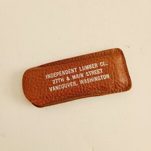 May include: A brown leather money clip with the text "INDEPENDENT LUMBER CO. 27TH & MAIN STREET VANCOUVER, WASHINGTON" printed in white. The clip is rectangular with rounded edges and a textured surface.