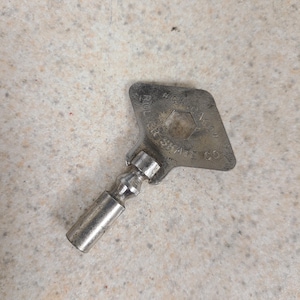 May include: A silver-toned metal skate key featuring a diamond-shaped handle with a central hexagonal cutout. The handle is embossed with the text CHICAGO ROLLER SKATE CO. The tool has a cylindrical shaft with a rounded, stepped neck design.