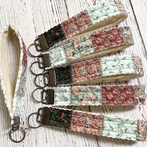 May include: Five fabric wristlet keychains featuring a quilted patchwork design with floral patterns in shades of sage green, dark green, and dusty rose. Each keychain includes a bronze-toned metal clasp and a circular split ring for keys.