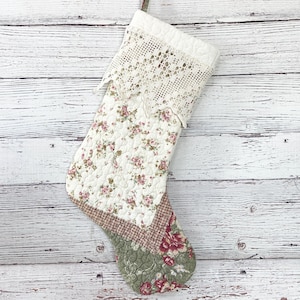 May include: A white quilted Christmas stocking with a floral pattern and a crocheted lace trim. The stocking is decorated with red and green patchwork fabric.