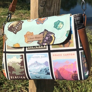 May include: A light blue and black crossbody bag with a brown strap. The bag is decorated with colorful patches featuring national park names and logos, including "Glacier Bay National Park" and "Grand Canyon National Park". The bag has a brown leather tag with the text "Aunt Henri" on it.