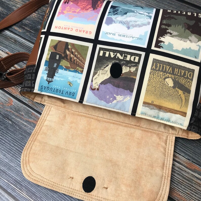May include: A brown leather crossbody bag with a flap closure. The bag is lined with a beige fabric and has a black fabric exterior with a collage of vintage travel posters featuring national parks like Glacier, Grand Canyon, Denali, and Death Valley.