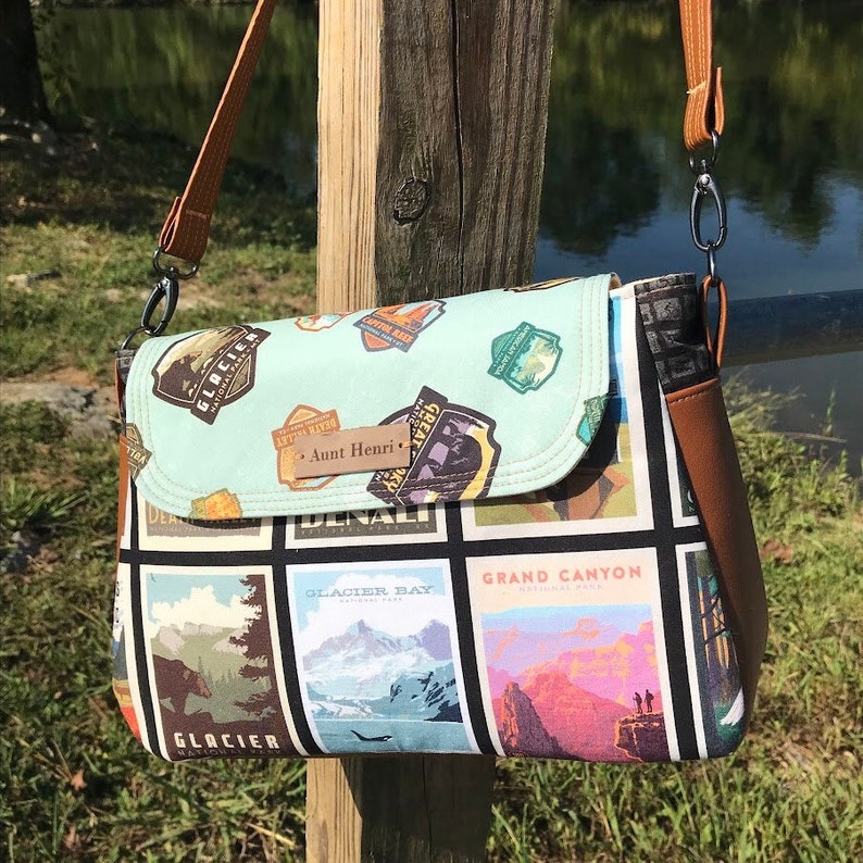 May include: A light blue and black crossbody bag with a brown strap. The bag is decorated with colorful patches featuring national park names and logos, including "Glacier Bay National Park" and "Grand Canyon National Park". The bag has a brown leather tag with the text "Aunt Henri" on it.