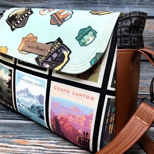 May include: A brown and black crossbody bag with a flap closure. The bag is made of canvas and features a patchwork design of national park logos and images. The strap is adjustable and has a metal clasp. The bag has a personalized tag that says "Aunt Henri".