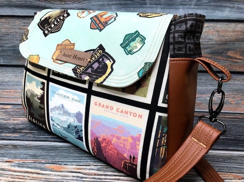 May include: A brown and black crossbody bag with a flap closure. The bag is made of canvas and features a patchwork design of national park logos and images. The strap is adjustable and has a metal clasp. The bag has a personalized tag that says "Aunt Henri".