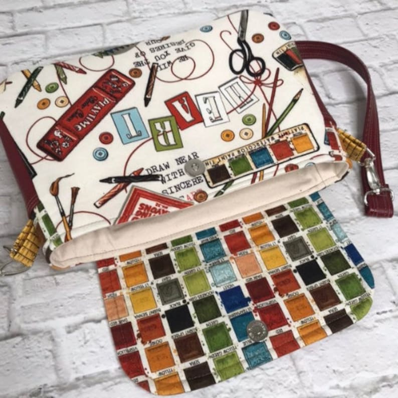 May include: A white fabric clutch bag with a red and white print of various art supplies, including paintbrushes, pencils, scissors, and thread. The bag has a red zipper and a red strap. The inside of the bag is lined with a fabric featuring a colorful grid of squares.