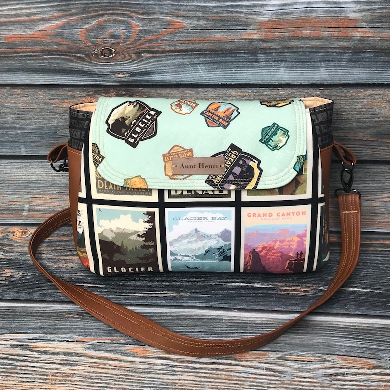 May include: A brown leather crossbody bag with a black and white fabric featuring images of national parks and the text "Aunt Henri".