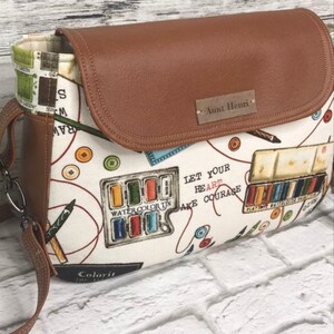 May include: A brown leather crossbody bag with a white fabric lining featuring a watercolor palette, pencils, and the text "Let your heart take courage". The bag has a flap closure with a brown leather tag that reads "Aunt Henri".