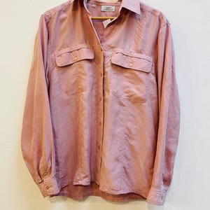 May include: A dusty rose button-down shirt with two chest pockets and long sleeves. The shirt is hanging on a wooden hanger against a white background. The fabric has a slight sheen.