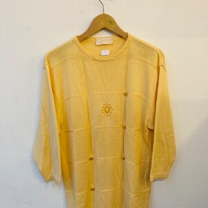Peut inclure: Un pull en maille jaune pâle avec un motif géométrique et des boutons dorés. Le pull a un col rond et des manches trois quarts. L'étiquette indique "Celiné".