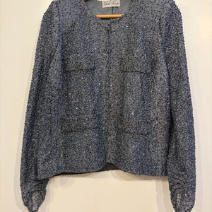 May include: A vintage, long-sleeved, blue sequined jacket with a round neckline. The jacket is fully covered in small, shimmering sequins and has a simple, button-less design. The jacket is hanging on a wooden hanger.