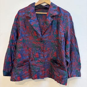 May include: A vintage blazer with a vibrant, abstract pattern in shades of purple, red, green, and blue. The jacket features a classic notched collar, three buttons, and two front pockets. It hangs on a wooden hanger against a plain white background.