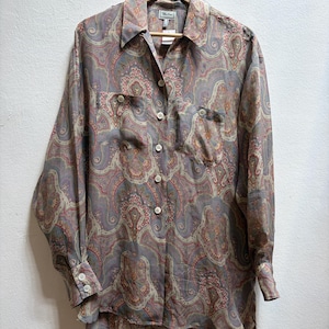May include: A long-sleeved button-down shirt with a paisley pattern in shades of gray, pink, and beige. The shirt has two chest pockets and is hanging on a wooden hanger.
