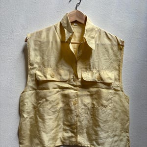May include: A sleeveless, pale yellow blouse with a collared neckline and two buttoned chest pockets. The blouse has a button-down front and is hanging on a wooden hanger. The fabric has a subtle sheen.