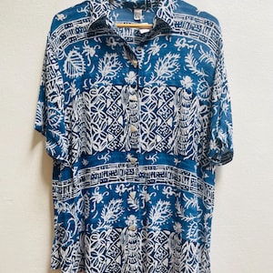 May include: A short-sleeved button-down shirt in a blue and white floral pattern. The shirt is hanging on a wooden hanger. The fabric features a repeating design of stylized leaves and geometric shapes.