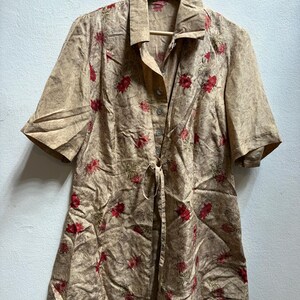 May include: A tan button-down shirt with short sleeves, featuring a floral pattern of red flowers. The shirt is hanging on a wooden hanger against a white wall. It has a tie at the waist.