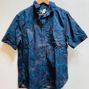 Op de afbeelding: Een shirt met korte mouwen met een bloemenpatroon in blauw, paars en bruin. Het shirt hangt aan een houten hanger. De kraag is open en het shirt heeft een voorzak. Het label luidt "MO PI".