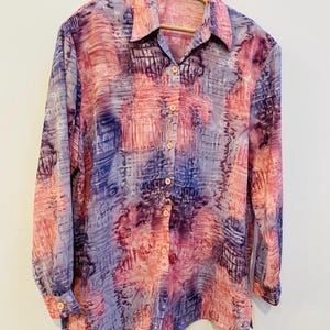 May include: A long-sleeved button-down shirt with a colorful abstract pattern. The shirt features a blend of pink, purple, and blue hues, creating a watercolor effect. It hangs on a wooden hanger against a white background.