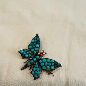 May include: A butterfly-shaped brooch with turquoise-colored beads and a red center stone. The brooch has a silver-colored metal frame and a pin closure. The wings are detailed with small, round beads.