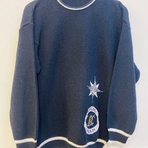 Peut inclure: Pull en maille bleu marine avec bordures blanches au col, aux poignets et à l'ourlet. Le pull présente un motif nautique avec une étoile et les mots "Sailing Team" brodés sur le devant. Le pull est suspendu à un cintre en bois.