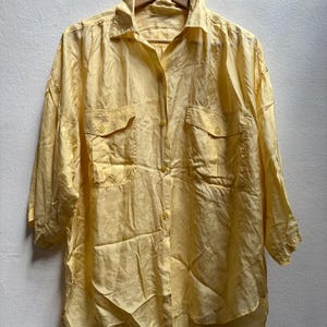 May include: A pale yellow button-down shirt with two chest pockets and three-quarter sleeves. The shirt is hanging on a wooden hanger against a white wall. The fabric has a subtle sheen.