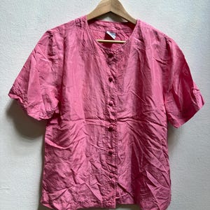 May include: A pink short-sleeved blouse with a round neckline and button-down front. The blouse is made of a lightweight, slightly wrinkled fabric and hangs on a wooden hanger. The sleeves are slightly puffed.