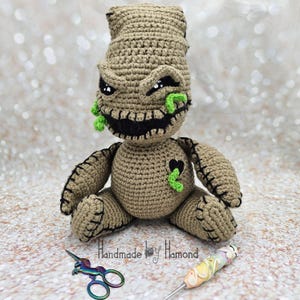 May include: A handmade crochet doll in a seated position, featuring a tan body with black stitched details. The doll has a menacing expression with a black mouth, white eyes, and green accents. The doll's design is inspired by a character from a popular animated film.