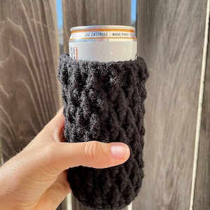 May include: Black crocheted can cosy with a white can inside. The can has the text "100 Calories - Made Pure" printed on it.