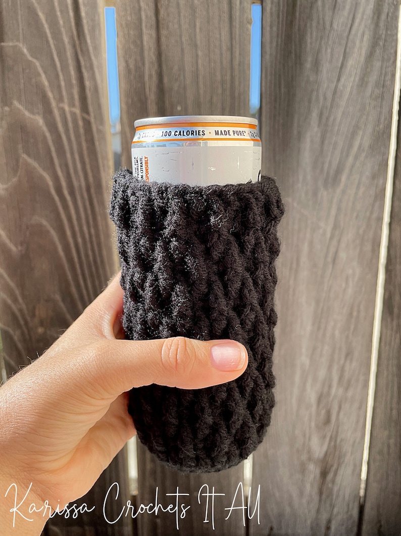 May include: Black crocheted can cosy with a white can inside. The can has the text "100 Calories - Made Pure" printed on it.