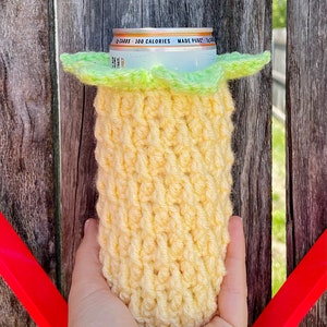 May include: A yellow crocheted can cooler with a green crocheted top. The cooler is holding a silver can with the text "2g CARBS - 100 CALORIES - MADE PURE" on it.