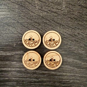 May include: Four wooden buttons with a geometric mountain design and a star pattern. The buttons are light brown and have two holes each.