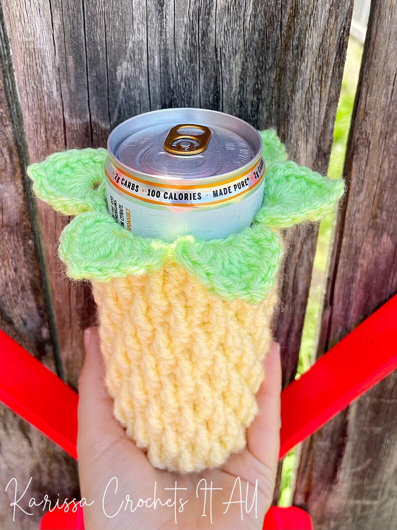 May include: A crocheted pineapple-shaped can cooler in yellow and green. The cooler is holding a silver can with the text "2g CARBS - 100 CALORIES - MADE PURE - 24 FL OZ" printed on it.