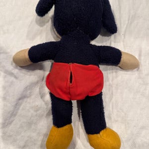 May include: A plush toy featuring a dark blue fuzzy body, long ears, and tan hands. The toy wears bright red shorts with a vertical opening in the center and has solid yellow feet. The character is shown from the back, displaying the texture of the fabric and the construction of the clothing.