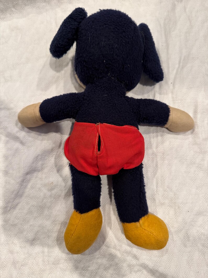 May include: A plush toy featuring a dark blue fuzzy body, long ears, and tan hands. The toy wears bright red shorts with a vertical opening in the center and has solid yellow feet. The character is shown from the back, displaying the texture of the fabric and the construction of the clothing.