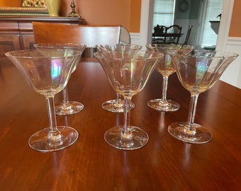 May include: Six clear, iridescent cocktail glasses with a flared bowl and long stem are arranged on a wooden table. The glasses have a subtle, shimmering effect, reflecting light with a rainbow sheen. The stem is thin and elegant.