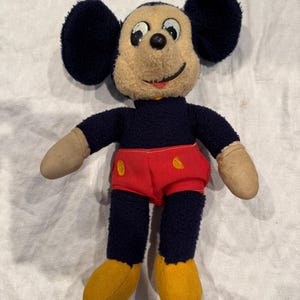 May include: A vintage plush toy featuring a character with large round black ears, a beige face, and a black nose. The figure wears a dark blue torso, bright red shorts with two yellow oval buttons, and solid yellow shoes with beige hands.