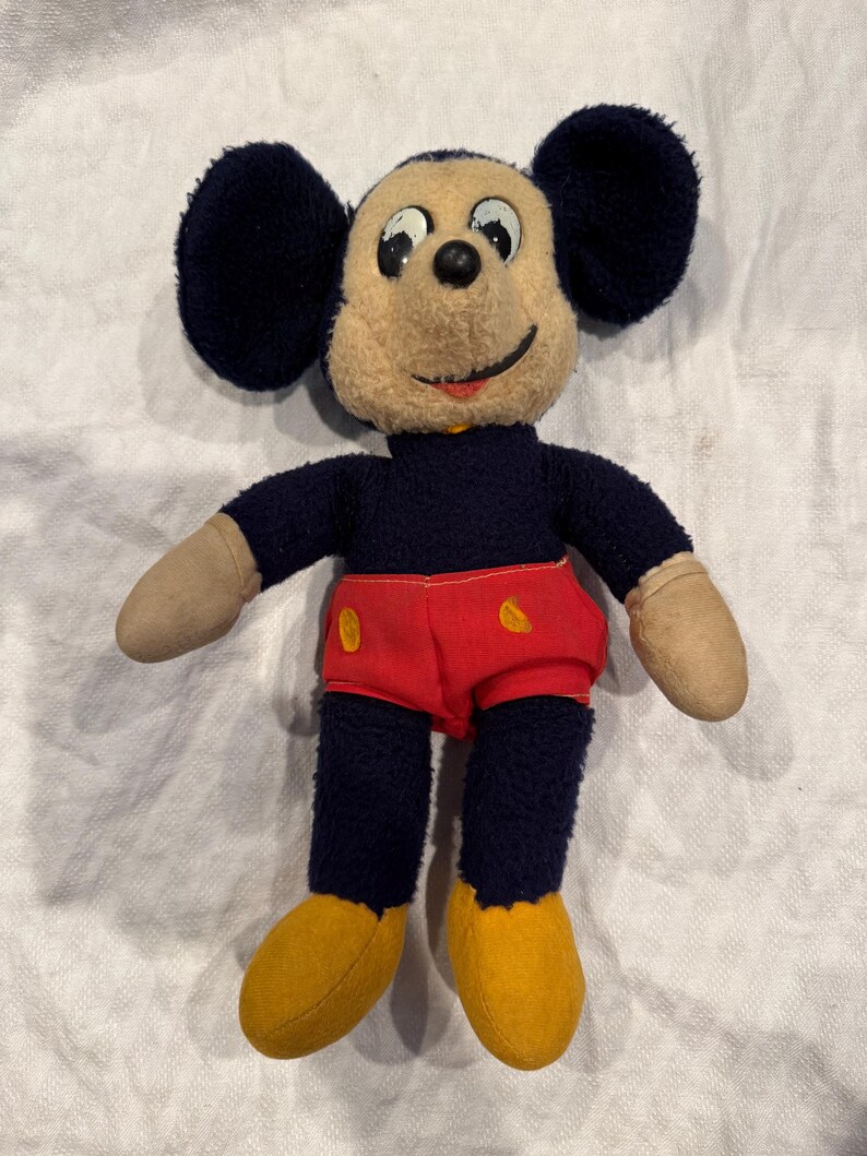 May include: A vintage plush toy featuring a character with large round black ears, a beige face, and a black nose. The figure wears a dark blue torso, bright red shorts with two yellow oval buttons, and solid yellow shoes with beige hands.