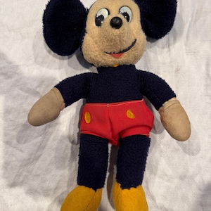 May include: A vintage plush toy featuring a character with large round black ears, a tan face, and a black body. The character wears bright red shorts with two yellow oval accents, tan gloves, and yellow shoes. The toy has a stitched smile.