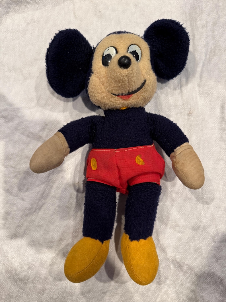 May include: A vintage plush toy featuring a character with large round black ears, a tan face, and a black body. The character wears bright red shorts with two yellow oval accents, tan gloves, and yellow shoes. The toy has a stitched smile.
